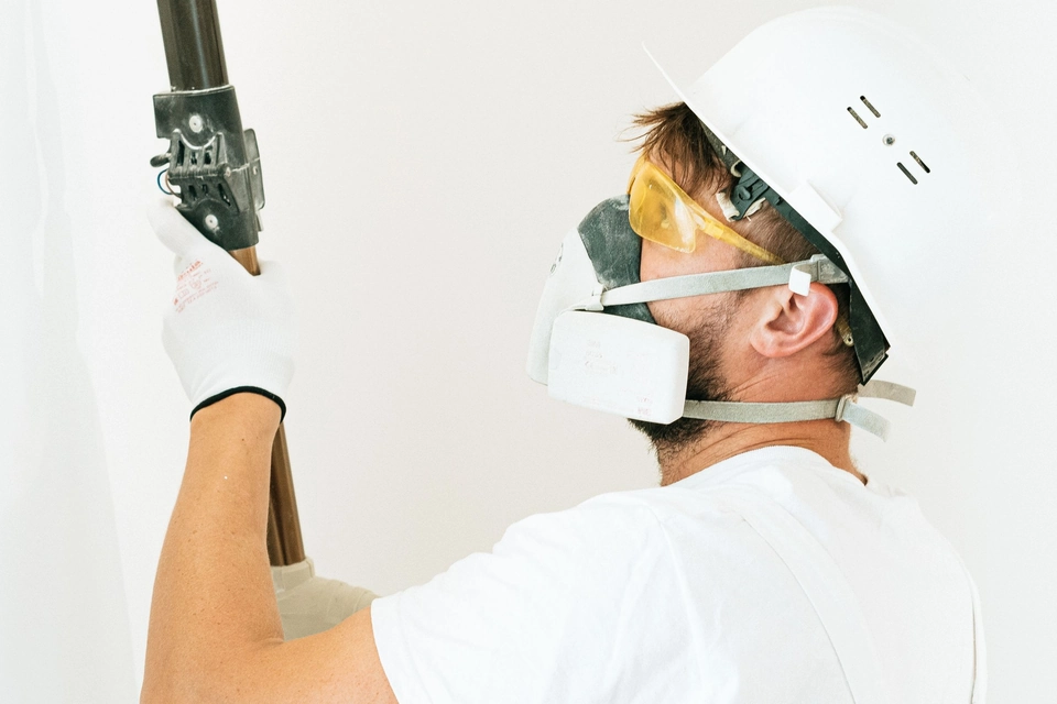 Schilder met helm en stofmasker werkt met een roller op een steel aan een lichte binnenwand